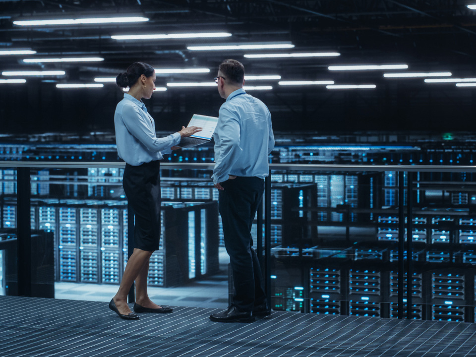 Two IT professionals stand on a walkway in a large, modern data center filled with server racks.