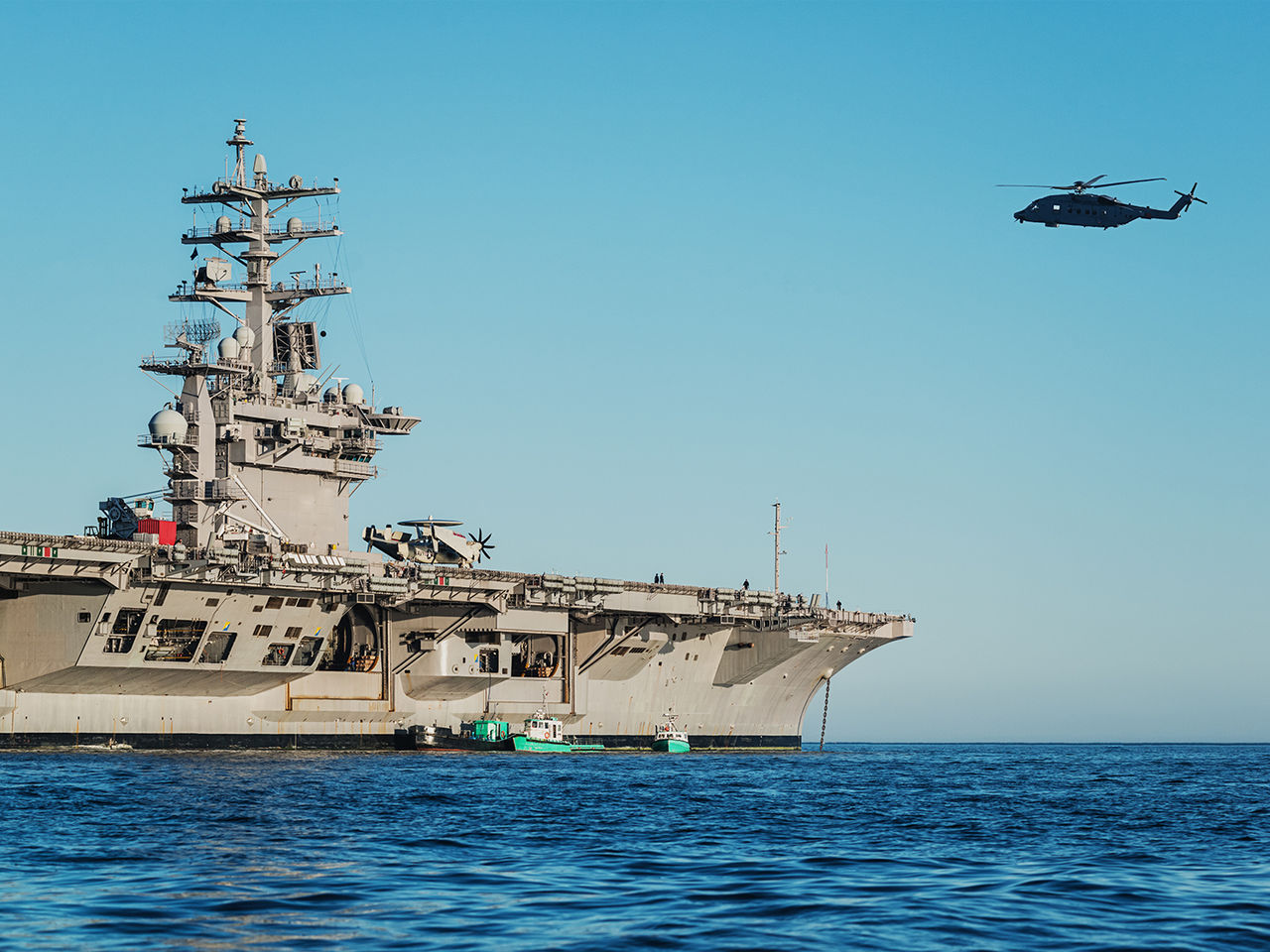 A US Navy aircraft carrier with a helicopter flying in the background.