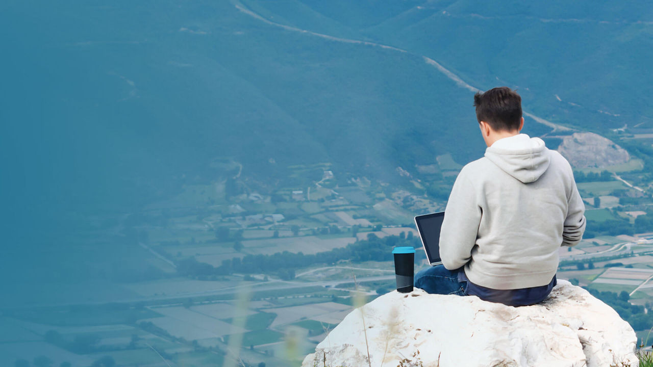 Working remotely on a laptop while sitting on a mountaintop, overlooking a vast landscape.